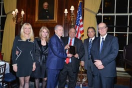 John Catsimatidis and an older man holding a plaque beside an American flag, with Margo Catsimatidis and three other guests