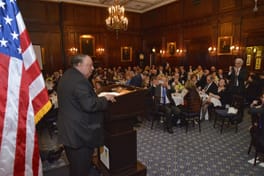 A speaker at the lectern beside an American flag, the dining room in view