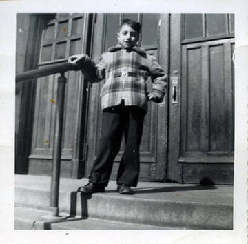 A black-and-white snapshot of John Catsimatidis as a boy, standing on a stoop in a plaid jacket, one hand on the railing
